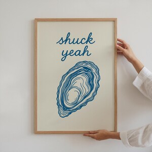 May include: A framed art print with the words "shuck yeah" in blue script above a blue oyster illustration. The artwork has a cream-colored background and a natural wood frame.