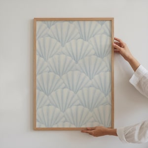 May include: A framed print with a repeating pattern of light blue seashell designs on a cream background. The print is in a natural wood frame. The artwork is held by a person wearing a white shirt.