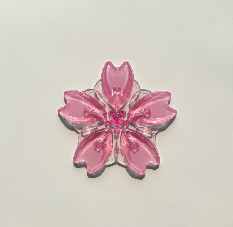 May include: A clear, pink, five-petal flower-shaped object with a small, pink center. The petals are a translucent pink, and the base is clear. The flower is centered on a white background.