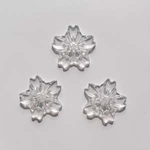 May include: Three clear plastic flower-shaped embellishments. Each flower has five petals and a center with a raised design.