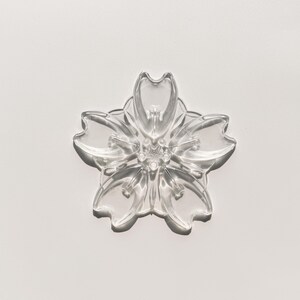 May include: A clear, five-petal flower-shaped object. The petals have rounded tips and a slightly textured surface. The center of the flower has a detailed, multi-layered design. The object is transparent, allowing light to pass through, and is set against a white background.