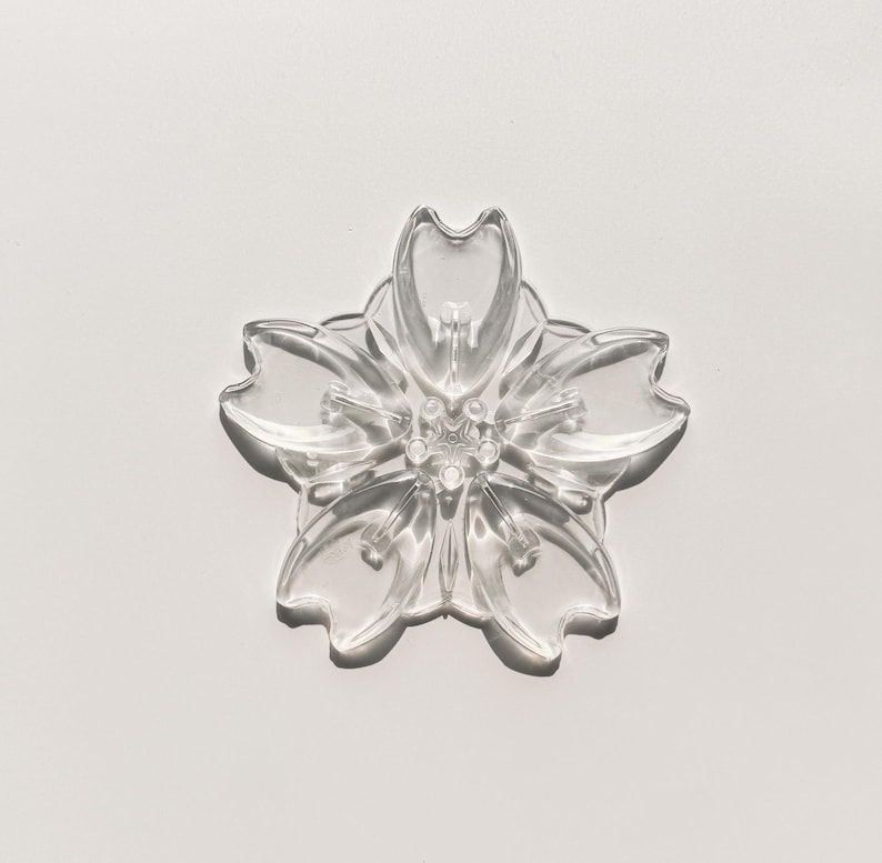 May include: A clear, five-petal flower-shaped object. The petals have rounded tips and a slightly textured surface. The center of the flower has a detailed, multi-layered design. The object is transparent, allowing light to pass through, and is set against a white background.