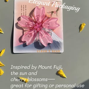 May include: A pink cherry blossom-shaped accessory on a card with text that reads "What's more? Elegant Packaging." The design is inspired by Mount Fuji and cherry blossoms. Yellow petals are scattered around.