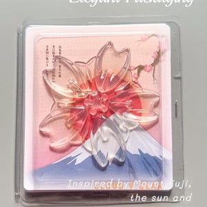 May include: Clear, transparent cherry blossom-shaped object with red accents, packaged in a clear plastic case. The packaging features a Mount Fuji illustration and text that reads "Inspired by Mount Fuji, the sun and cherry blossoms".