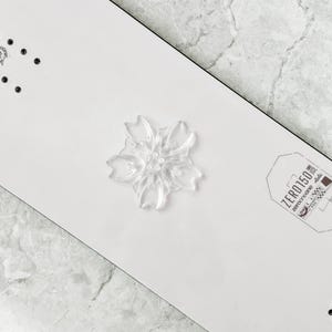 May include: A clear, flower-shaped decorative element is centered on a white surface. The flower has multiple petals and a detailed center. The surface has a black border and a small tag with text that reads "ZERO150".