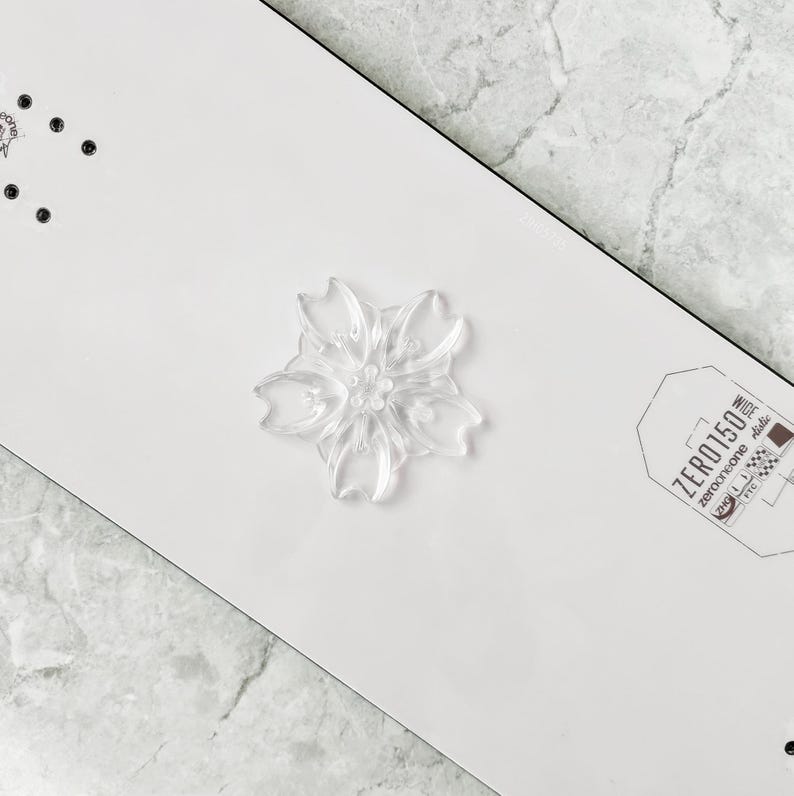 May include: A clear, flower-shaped decorative element is centered on a white surface. The flower has multiple petals and a detailed center. The surface has a black border and a small tag with text that reads "ZERO150".