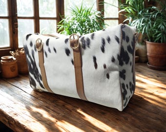 May include: A large, rectangular travel bag with a cowhide pattern of white and black spots. The bag features tan leather straps and gold-coloured metal hardware. The bag is on a wooden surface.