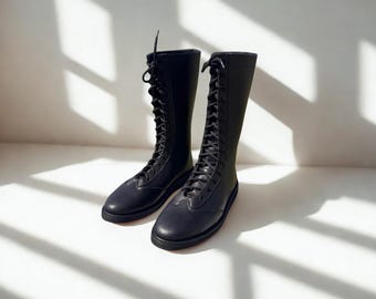 May include: A pair of black, knee-high boots with a lace-up design. The boots are made of a smooth material and have a flat sole. The boots are displayed on a white surface with shadows.