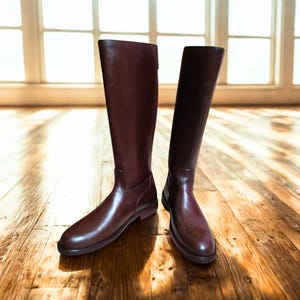 May include: A pair of tall, knee-high brown leather boots. The boots have a smooth, glossy finish and a classic design. They are standing on a wooden floor, with natural light coming from a window in the background.