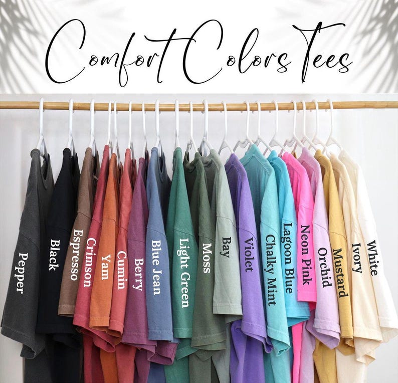May include: A row of 16 t-shirts hanging on a rack, showcasing a variety of colors, including white, ivory, mustard, orchid, neon pink, lagoon blue, chalky mint, violet, bay, moss, light green, blue jean, berry, cumin, yam, crimson, espresso, and black. The t-shirts are labeled with their respective colors.