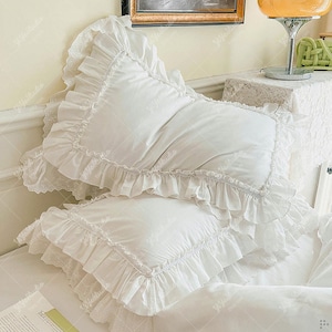 May include: Two white rectangular pillows with ruffled edges and lace detailing. The pillows are made of a soft, smooth fabric. The ruffles and lace trim give a delicate, feminine appearance, suitable for a bedroom setting.