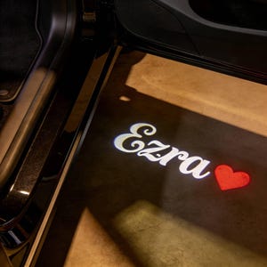 May include: A car door's interior is visible, with a light projecting the name "Ezra" in white script, accompanied by a red heart symbol. The light shines on a tan surface, creating a personalized effect.