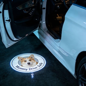 May include: A white car door is open, illuminating a circular light on the ground. The light features a corgi dog image and the text "Mommy Drive Safe". The car's interior is visible.
