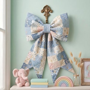 May include: A large, decorative fabric bow featuring a patchwork design of light blue, white, and soft pink floral and gingham patterns. The bow is quilted with intricate stitching and hangs from a decorative gold-toned metal wall hook.