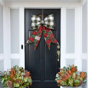 May include: A large decorative bow featuring a top section with a green and white buffalo check pattern and two long, pointed tails decorated with a red and green poinsettia floral print, hanging on a solid black front door.