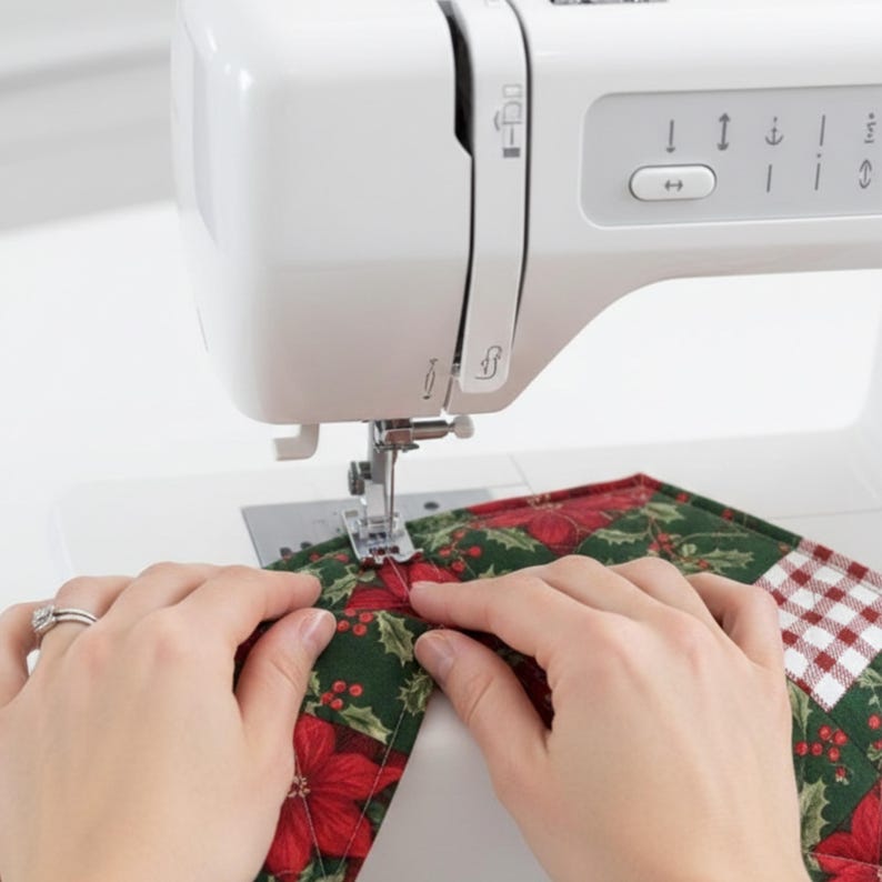 May include: A white sewing machine with a needle and presser foot stitching a piece of fabric featuring a festive pattern of red poinsettias, green holly leaves, and a red and white checkered square design, positioned under the machine needle.
