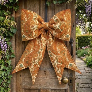 May include: A large bow made from paisley fabric in orange, brown, and cream tones, is attached to a weathered wooden door. The bow's detailed pattern and warm colours create a welcoming, rustic aesthetic, ideal for seasonal decoration.