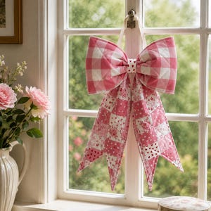 May include: A quilted fabric bow decoration featuring a pink and white gingham pattern on the top loops and a patchwork design on the two long tails. The patchwork consists of various pink and white floral and heart-shaped prints on fabric.