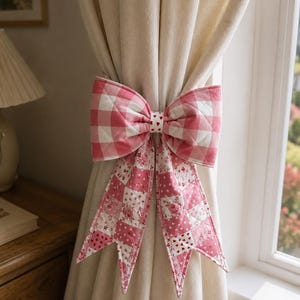 May include: A quilted fabric curtain tieback featuring a large bow with a pink and white gingham pattern. Two long, pointed tails hang down, showcasing a patchwork design of pink floral prints and small red hearts on a white background.