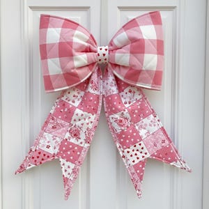 May include: A large, decorative bow in shades of pink and white, perfect for door decor. The bow features a quilted gingham pattern on the upper portion and a patchwork design with hearts and floral prints on the tails. The bow is made of fabric.