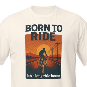 May include: Cream-colored t-shirt with a graphic of a cyclist on a road at sunset. The text "BORN TO RIDE" is above the image, and "It's a long ride home" is below. The design features a silhouette of a cyclist.