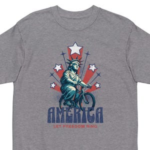 May include: A gray t-shirt featuring a graphic of the Statue of Liberty riding a bicycle, with red and white starbursts and stars. The word "AMERICA" is printed in blue, with "LET FREEDOM RING" below.