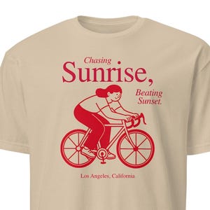 May include: A beige t-shirt featuring a red illustration of a person cycling. The text above reads "Chasing Sunrise, Beating Sunset", with "Los Angeles, California" printed below the graphic.