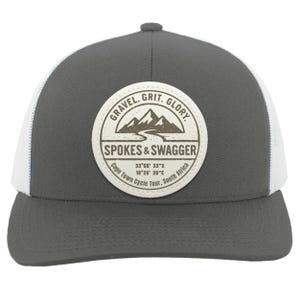 May include: A gray trucker hat with a white mesh back and a curved brim. The hat features a circular patch with the text "GRAVEL. GRIT. GLORY" and "SPOKES & SWAGGER". Coordinates for Cape Town Cycle Tour, South Africa are also printed on the patch.