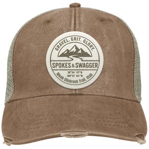 May include: A brown trucker hat with a distressed finish. The hat features a circular patch with the text "GRAVEL. GRIT. GLORY." and "SPOKES & SWAGGER." The patch also includes coordinates and "Moab Slickrock Trail, Utah."