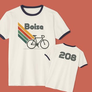 May include: Cream-colored t-shirt with navy blue trim featuring the word "Boise" above a bicycle graphic and a retro color stripe design. The back of the shirt displays the number "208" in a matching font.