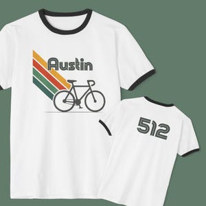 May include: White t-shirts with black trim. One shirt features the word "Austin" with a bicycle graphic and a retro color scheme. The other shirt displays the number "512" in a bold font.