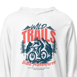 May include: White long-sleeve hooded shirt with a graphic of a mountain biker. The design features the words "WILD TRAILS" in red and a mountain scene with trees. Below the graphic, it says "Bold Adventures".