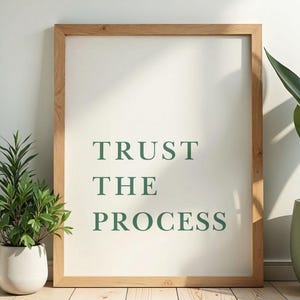 May include: A framed print with the words "TRUST THE PROCESS" in green lettering. The frame is made of light-colored wood. A potted plant is on the left, and another is on the right. The background is a light-colored wall and wooden floor.