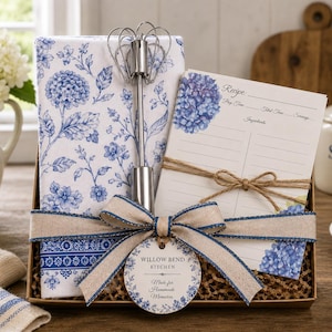 May include: A kitchen gift set containing a white towel with a blue floral pattern, a silver metal whisk, and a recipe card with blue hydrangea illustrations. The items are tied with a tan ribbon and a round tag labeled Willow Bend Kitchen.
