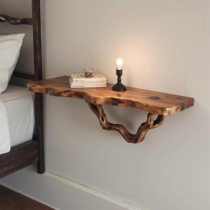 Puede incluir: Mesa de noche flotante de madera con borde natural. La mesa es de madera natural y tiene una forma orgánica única. Una lámpara negra y libros están colocados sobre la mesa. La mesa está montada en la pared. La mesa mide unos 100 cm de largo.