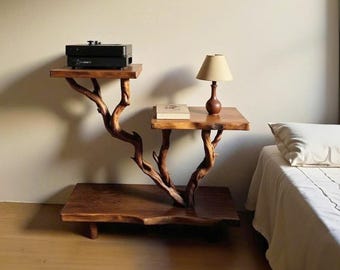 May include: A unique, handcrafted wooden side table with a natural tree branch design. The table features three platforms, one holding a black electronic device, another a book, and the top platform supporting a small lamp. The wood has a warm, natural finish.