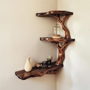 May include: A wooden shelf unit shaped like a tree with three shelves. The shelves are made of natural wood and have a rustic finish. The shelf unit is mounted on a wall and is decorated with a vase, a glass jar, and other decorative items.