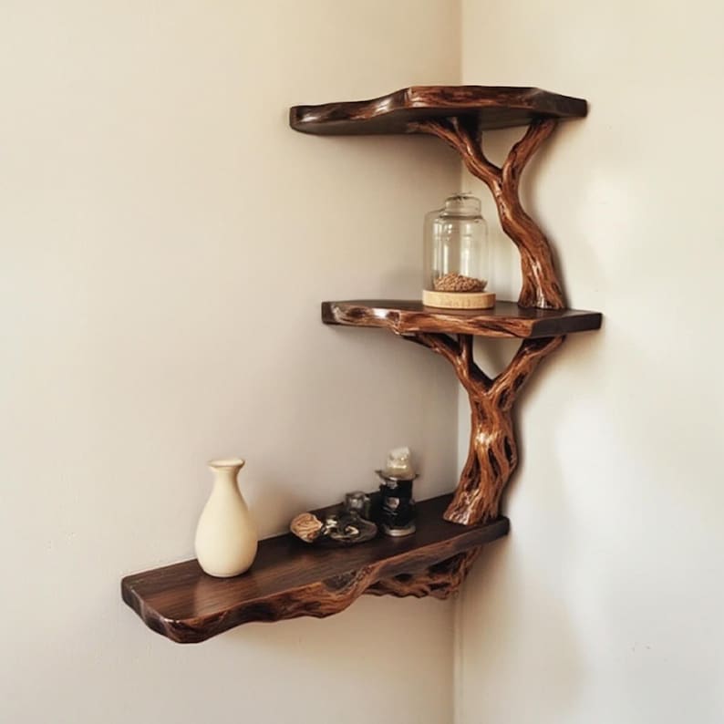 May include: A wooden shelf unit shaped like a tree with three shelves. The shelves are made of natural wood and have a rustic finish. The shelf unit is mounted on a wall and is decorated with a vase, a glass jar, and other decorative items.