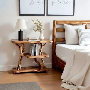 May include: A rustic, wooden nightstand with a two-tiered design. The nightstand features a natural wood finish and is supported by tree branch-like legs. A black lamp, books, and a vase with greenery are on top. The nightstand is next to a bed.