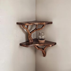 May include: A two-tiered corner shelf made from natural wood branches. The shelf is stained a dark brown color and has a rustic, organic look. A small glass jar with black contents sits on the top shelf.