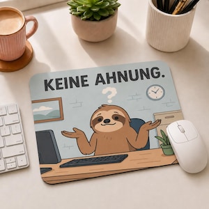 Könnte beinhalten: Ein rechteckiges Mauspad mit einer Cartoon-Illustration eines braunen Faultiers, das an einem Schreibtisch sitzt und die Arme schulterzuckend ausbreitet. Über dem Faultier steht der Text "KEINE AHNUNG.". Das Bild zeigt einen Schreibtisch, Computer und Uhr.