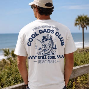 Op de afbeelding: Crèmekleurig T-shirt met een marineblauwe print op de rug. De print toont een bas met een zonnebril en vissershoed, met de tekst "COOL DADS CLUB" en "STILL COOL - JUST TIRED". Het T-shirt heeft korte mouwen.