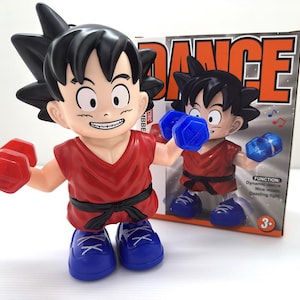 Op de afbeelding: Een speelgoedfiguur van een personage met stekelig zwart haar, gekleed in een rood shirt, zwarte riem en blauwe schoenen, met rode gewichten. De figuur staat voor een doos met het woord "DANCE".