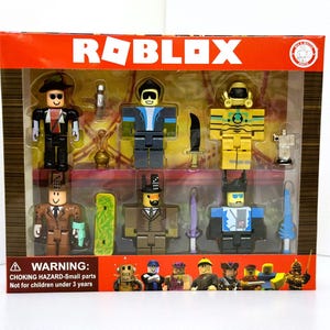 Könnte beinhalten: Eine Box mit Roblox-Actionfiguren. Die Figuren sind in verschiedenen Farben und Outfits gehalten, mit Zubehör wie Schwertern und Hüten. Die Verpackung ist orange und zeigt das Roblox-Logo sowie einen Warnhinweis auf Kleinteile.