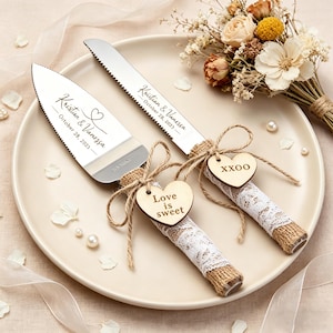 Puede incluir: Un juego de servir pastel de bodas sobre un plato. El juego incluye un cuchillo y un servidor de pastel con texto personalizado. Los mangos están envueltos con arpillera y encaje, y adornados con etiquetas de corazón de madera. Un pequeño ramo y elementos decorativos completan la escena.