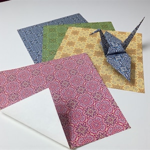 May include: Origami paper in blue, green, yellow, and pink with a floral pattern. A folded origami crane sits on a yellow sheet. The paper is square and has a white backing. The paper is approximately 15 cm x 15 cm.