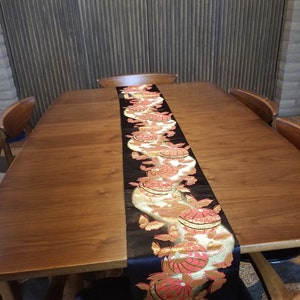 May include: A wooden dining table with a black and gold table runner featuring a repeating pattern of pink and gold circles and butterflies. The table runner is approximately 12 inches wide and 72 inches long.