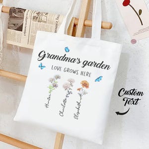 Puede incluir: Bolso de mano blanco con el texto "Grandma's garden LOVE GROWS HERE" e ilustraciones florales. Los nombres "Hunter", "Charlotte" y "Elizabeth" están escritos debajo de las flores. Mariposas azules dispersas.