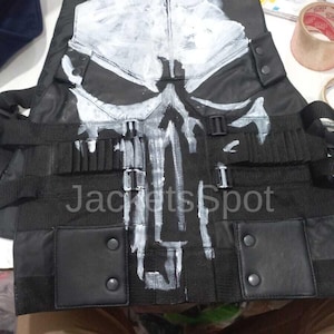 Op de afbeelding: Zwart tactisch vest met een wit schedelontwerp op de voorkant. Het vest heeft meerdere banden, zakken en de tekst "Jackets Spot" erop gedrukt. Het vest is waarschijnlijk voor cosplay of kostuums.