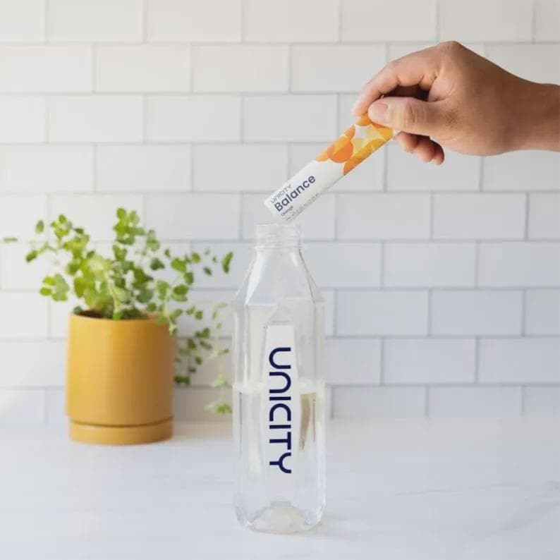 K&ouml;nnte beinhalten: Eine durchsichtige Kunststoff-Wasserflasche mit dem vertikal aufgedruckten blauen Unicity-Logo. Eine Hand h&auml;lt einen wei&szlig;en Unicity Balance Orange Nahrungserg&auml;nzungsmittel-Stick &uuml;ber die Flaschen&ouml;ffnung, um den Inhalt in das Wasser zu geben.